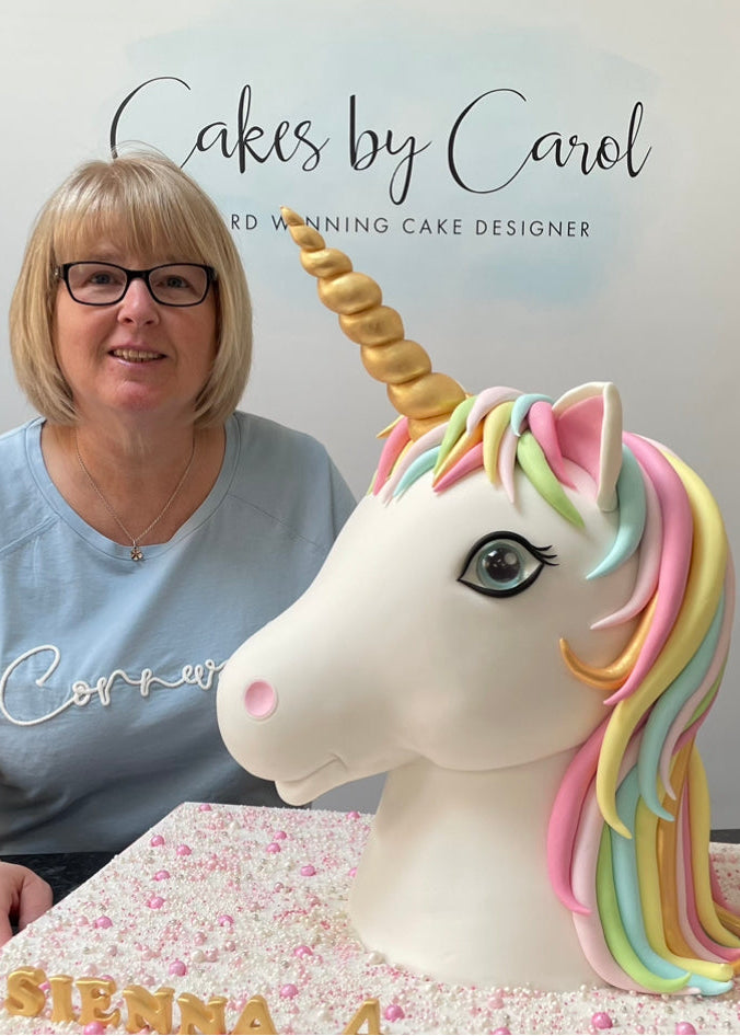 Person sitting next to a unicorn cake with 'Cakes by Carol' sign in the background