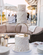 A white three layer cake with subtle petal designs on each layer, the top two tiers are separated by a Hoop Tier Separator - Prop Options